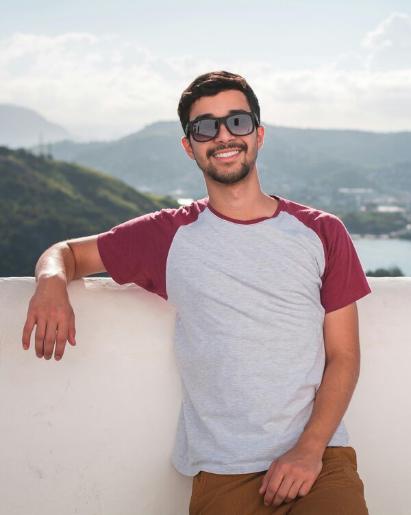 A smiling man with sunglasses on, in front of a white wall, with a scenic mountain background.